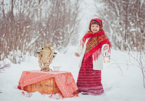 Adorable little girl in old clothes in a winter forest with a samovar and bagels