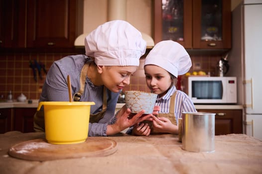 Happy family, loving mother and her little daughter, both of them wearing white chef's hat and beige apron, standing by kitchen island, sniffing dough while preparing cake together for Easter holiday