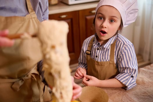 Amazed adorable Caucasian 5-6 yeras old child, a lovely little kid girl looking at her mom kneading dough while preparing delicious homemade cakes at home kitchen for Easter or Christmas holidays