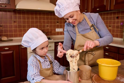 Loving cheerful mother and her adorable 5 years old daughter in chef's hats and beige aprons, enjoying cooking together, having fun, kneading dough while preparing homemade cakes for Easter holidays
