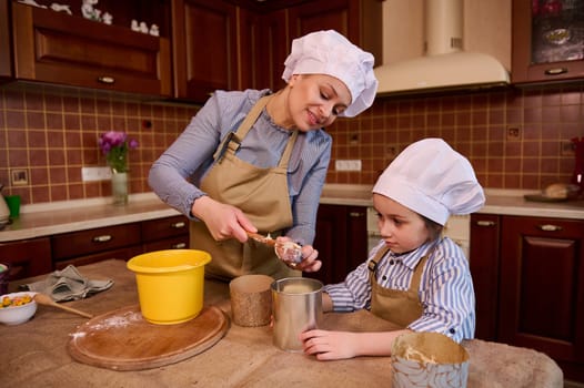 Authentic smiling happy woman, mother, confectioner removes raw dough from bowl and puts it into a baking dish, standing by kitchen table with her daughter, enjoying cooking Easter or Christmas cakes
