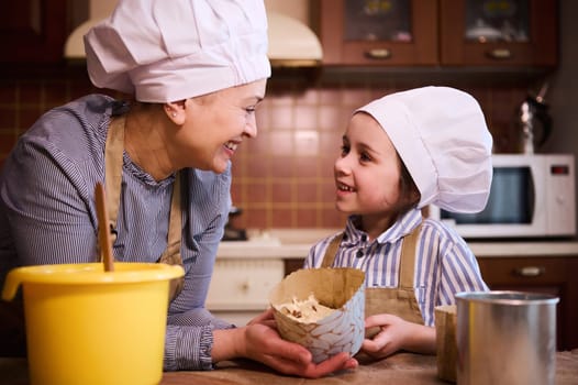 Happy loving mother and lovely child, adorable daughter dressed in chef's uniform, smiling looking at each other, holding a paper baking dish with raw dough while preparing Easter cake panettone