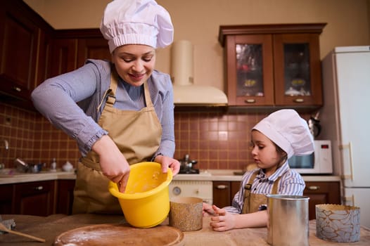 Happy middle-aged multi-ethnic woman, a loving mother and her cute daughter, adorable little child girl cooking together a panettone cake for the Easter table in the cozy wooden home kitchen interior