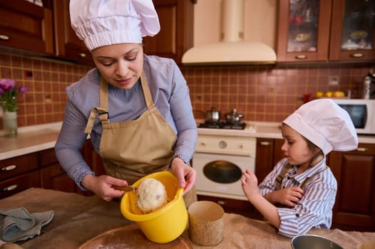 Selective focus on a multi-ethnic middle-aged pretty woman, mother dressed in beige chef's apron and hat, teaching her lovely daughter cooking, kneading dough while preparing festive cake for Easter