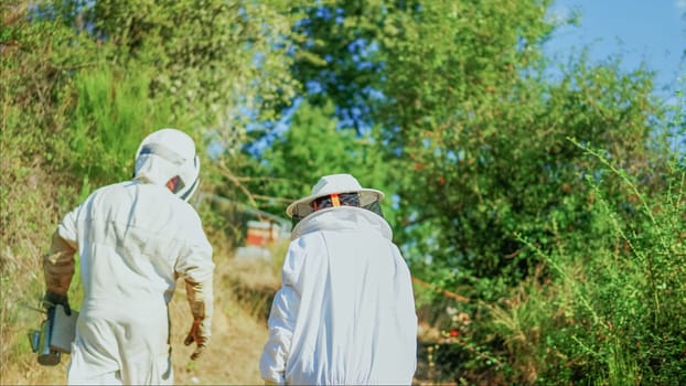 Two beekeepers with protective clothes walking towards the apiary in a sunny day