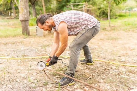 Man cutting a piece of iron using a radial saw in a yard with trees