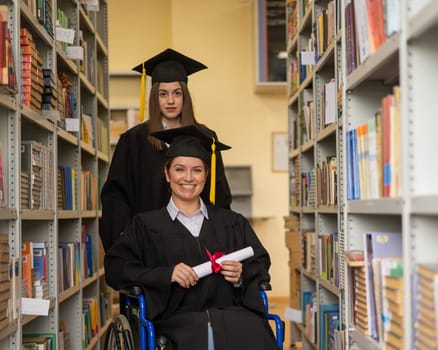 Happy young woman and woman in wheelchair in graduate gown with diploma in hands in library. Inclusive education