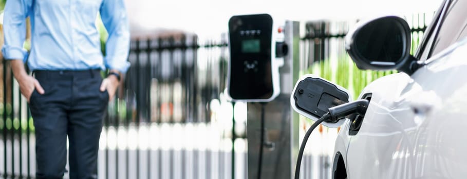 Closeup progressive suit-clad businessman with his electric vehicle recharge his car on public charging station in modern city with power cable plug and renewable energy-powered electric vehicle.