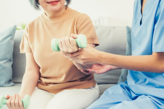 Caregiver helping contented senior woman exercise with dumbbell at home. Rehab for elder, senior medical care.