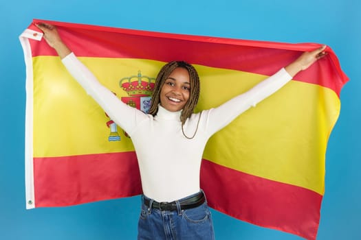 Studio portrait with blue background of a happy african woman raising an spanish flag