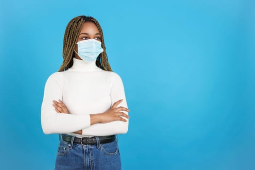 Studio portrait with blue background of a african woman standing with arms crossed wearing a facial protective mask