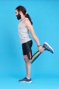 Vertical studio portrait with blue background of the profile of a man with a prosthesis on his leg stretching his quadriceps