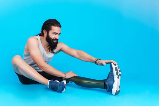 Studio photo with copy space and blue background of a sportive man with a leg prosthesis sitting on the ground stretching the femoral muscle.