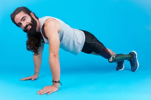 Horizontal studio portrait with blue background of a sportive man with a prosthetic leg doing push-ups on the floor