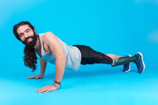 Horizontal studio portrait with blue background of a smiley and sportive man with a prosthetic leg doing push-ups on the floor