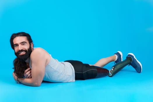 Horizontal studio portrait with blue background of a smiley and sportive man with a prosthetic leg lying on the floor
