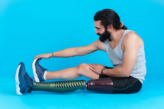 Studio photo with blue background of a sportive man with a leg prosthesis sitting on the ground stretching the femoral muscle.