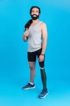Vertical Studio portrait with blue background of a man with a prosthesis on a leg, looking at the camera smiling and gesturing to be well with his thumb