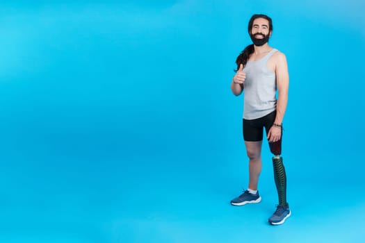 Studio portrait with blue background and copy space of a sportive man with a prosthesis on a leg smiling at the camera and gesturing to be well with his thumb