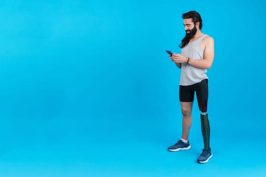 Studio portrait with blue background and copy space of a sportive man with a leg prosthesis using a mobile