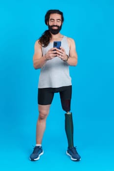 Vertical studio portrait with blue background of a distracted sportive man with a leg prosthesis using a mobile