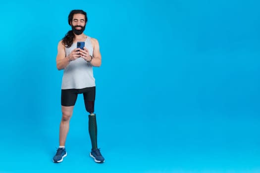 Studio portrait with blue background and copy space of a man with a prosthetic leg using a mobile phone
