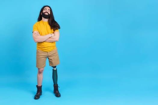 Studio portrait with copy space and blue background of a cheerful man with a leg prosthesis standing with arms crossed