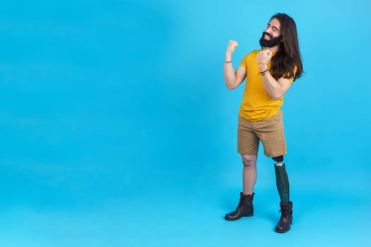 Studio portrait with blue background and copy space of a happy man with a leg prosthesis celebrating raising the fists
