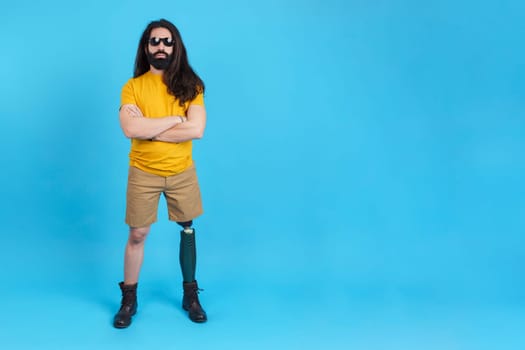 Studio portrait with blue background and copy space of a serious man with sunglasses standing with arms crossed and a prosthetic leg