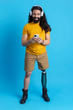 Studio portrait with blue background and copy space of a man with prosthetic leg standing using the mobile and listening to music