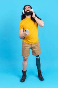 Vertical studio portrait with blue background of a man with prosthetic leg enjoying while listening to music with the mobile