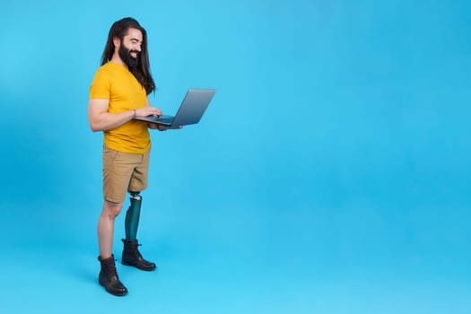 Studio portrait with blue background and copy space of a man with prosthetic leg standing and smiling using a laptop