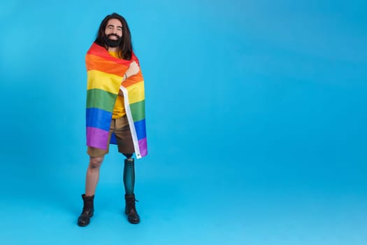 Studio portrait with blue background of a proud man with a prosthetic leg wrapped in a lgbt rainbow flag