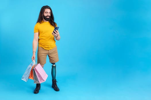 Studio portrait with blue background of a cool man with prosthetic leg using the mobile holing shopping bags