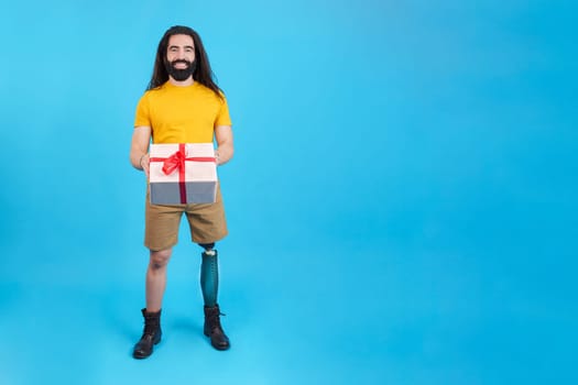 Studio portrait with blue background and copy space of a man with a leg prosthesis delivering a gift