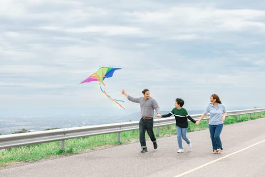 Progressive happy family vacation and carefree day concept. Young parents mother father and son run along and flying kite together road with enjoy natural scenic on scenery and clear sky background.