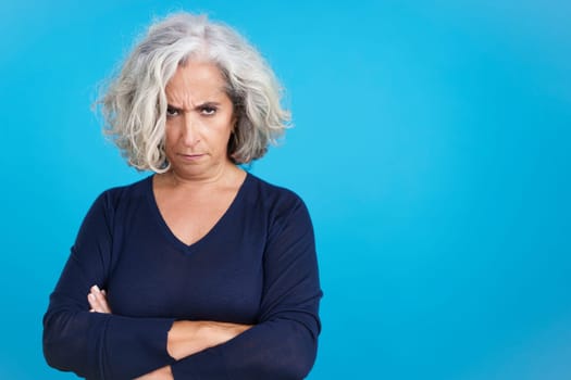 Studio portrait with blue background of a mature woman crossing the arms while looking at camera with an angry expression
