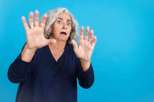 Studio portrait with blue background of a mature woman gesturing with her hands in panic