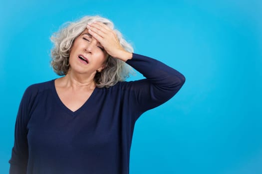 Studio portrait with blue background of a mature woman with hand on forehead expressing headache