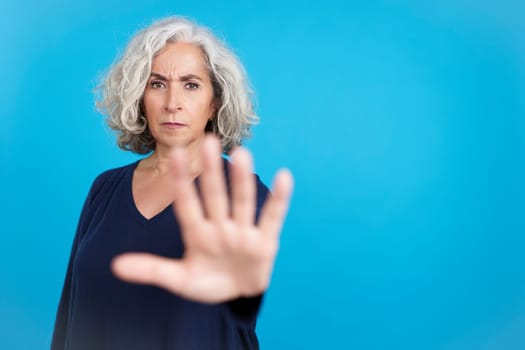Studio portrait with blue background of a serious mature woman gesturing with her hand prohibition and stop