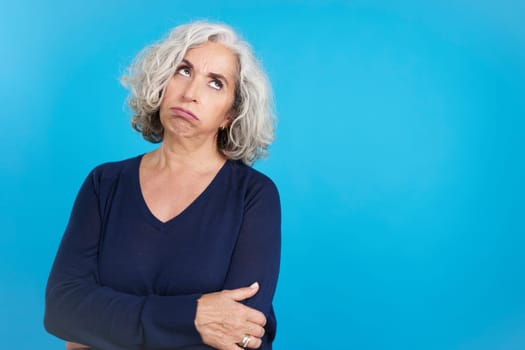 Studio portrait with blue background of a mature woman with arms crossed looking up with expression of boredom