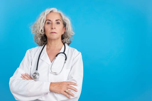 Studio portrait with blue background of a mature female doctor in white lab coat with crossed arms looking seriously at camera