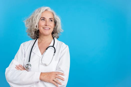 Studio portrait with blue background of a Mature female doctor in white coat looking up with the arms crossed