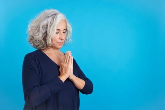 Studio portrait with blue background of a mature woman praying with folded hands and closed eyes