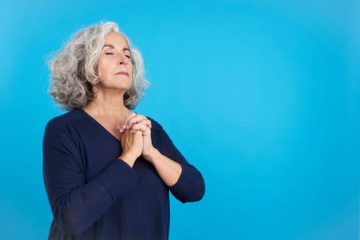 Studio portrait with blue background of a concentrated aged woman praying with the eyes closed