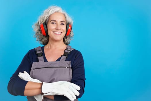 Studio portrait with blue background of an aged female carpentry worker with tools and work uniform smiling at the camera with arms crossed