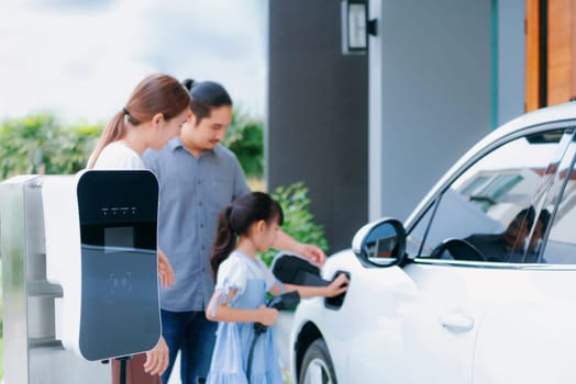 Closeup focus hand insert EV charger plug to electric vehicle at home charging station with blurred family in background. Progressive concept of green and clean energy to reduce CO2 emission by EV car