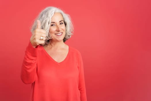 Studio portrait with red background of a mature woman gesturing agreement raising the thumb