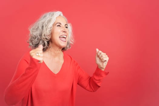Studio portrait with red background of an excited mature woman celebrating while raising the fists