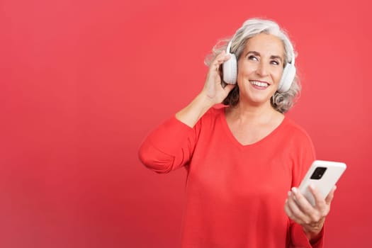 Studio portrait with red background of a distracted woman listening a podcast using the mobile and headphones